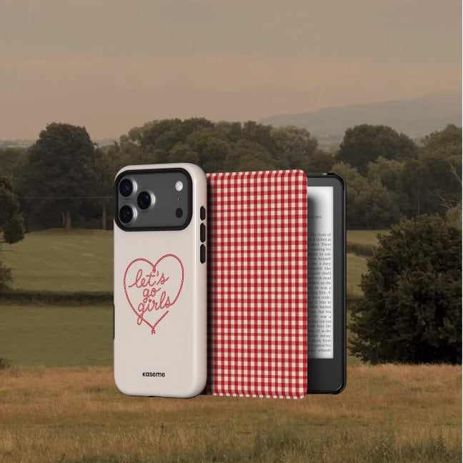 A white phone case with a red heart outline and 'let's go girls' text, shown next to a red-and-white gingham folio case and a black e-reader, set against an outdoor rural landscape background.