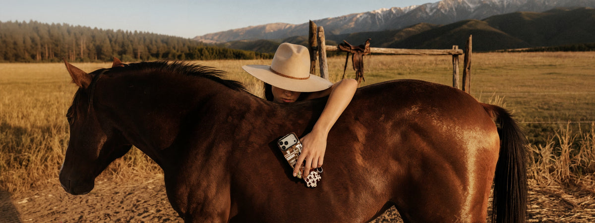 A person in a wide-brimmed hat leans against a brown horse in an outdoor ranch setting with mountains and a fence in the background. The person is holding a patterned phone case from the new collection Rodeo Muse.