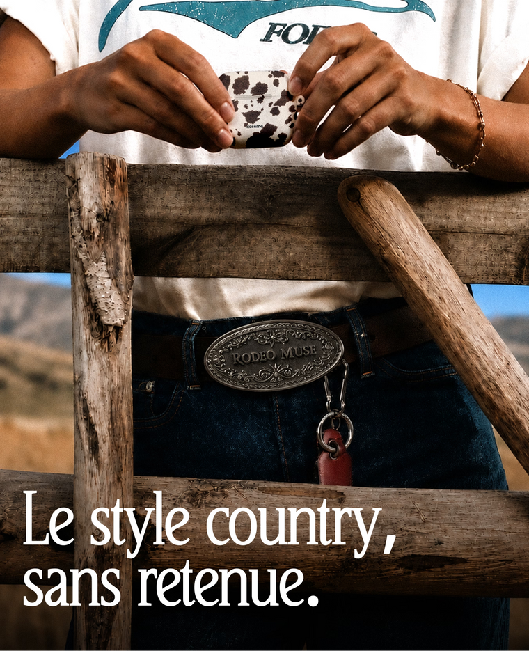 A person outdoors wearing a white graphic T-shirt, dark blue jeans, and a brown belt with a large engraved silver buckle that reads 'RODEO MUSE'. They are holding a small cow-print cup behind a rustic wooden fence, with a French text overlay at the bottom.
