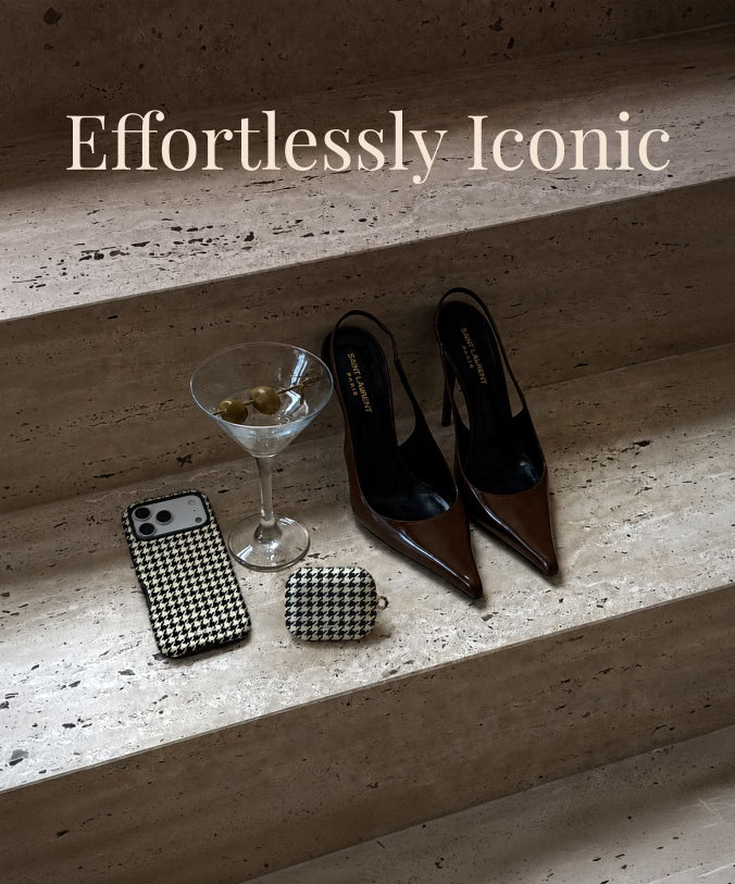 A styled flat-lay on stone steps featuring a pair of pointed-toe slingback heels in glossy brown and black, alongside a black-and-white houndstooth phone case, a matching AirPods Cases, a martini glass, and a smartphone. The text 'Effortlessly Iconic' appears at the top.