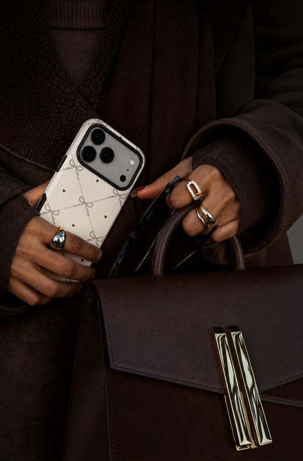 A close-up lifestyle shot of a person holding a white and black Gucci phone case, wearing gold rings, and holding a dark brown leather handbag with a gold-tone clasp.